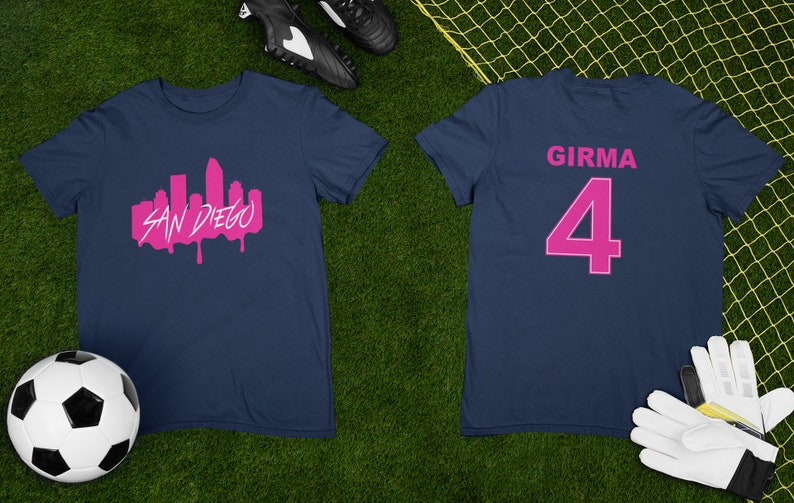 May include: Two navy blue t-shirts with pink lettering. The front of one shirt has a pink silhouette of the San Diego skyline with the words "SAN DIEGO" underneath. The back of the other shirt has the name "GIRMA" and the number "4" in pink.