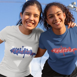 Puede incluir: Dos mujeres con camisetas con un gr&aacute;fico del horizonte de Seattle. Las camisetas son blancas y azules con texto rojo. Las mujeres sonr&iacute;en y miran a la c&aacute;mara.