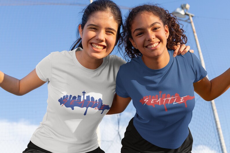 Puede incluir: Dos mujeres con camisetas con un gr&aacute;fico del horizonte de Seattle. Las camisetas son blancas y azules con texto rojo. Las mujeres sonr&iacute;en y miran a la c&aacute;mara.