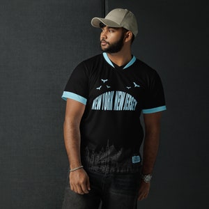 K&ouml;nnte beinhalten: Schwarzes T-Shirt mit einem hellblauen V-Ausschnitt und hellblauen Abschl&uuml;ssen an den &Auml;rmeln. Das Shirt hat einen grafischen Druck einer Stadtlandschaft mit dem Text "NEW YORK NEW JERSEY" und Flederm&auml;usen, die &uuml;ber der Stadt fliegen.