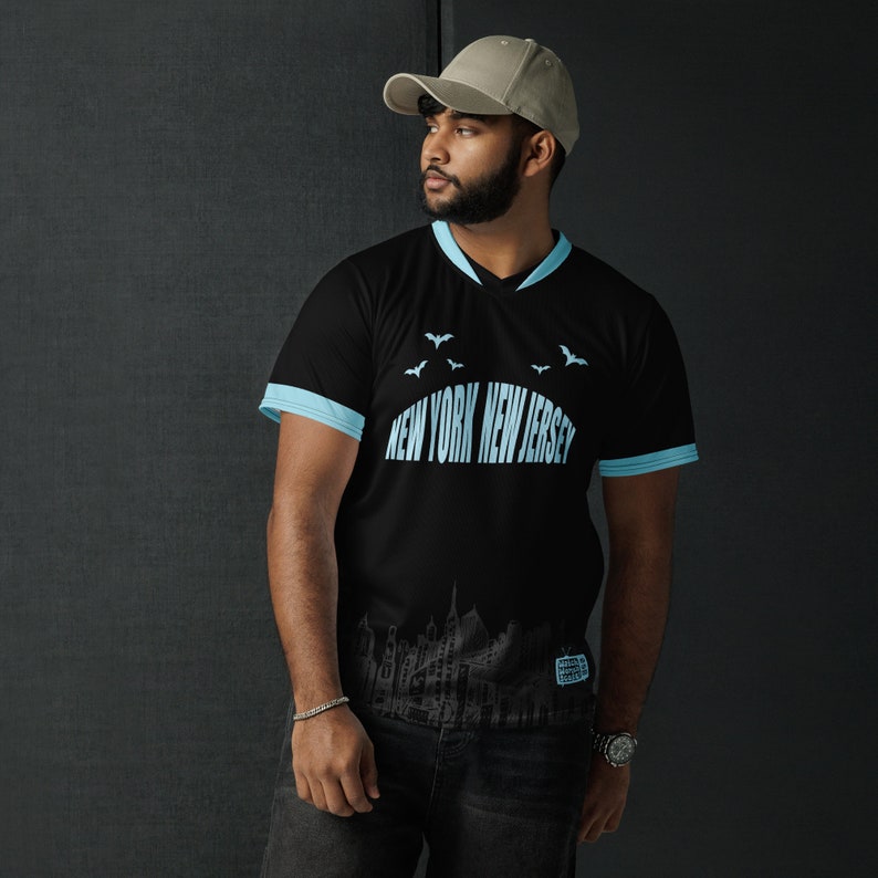 K&ouml;nnte beinhalten: Schwarzes T-Shirt mit einem hellblauen V-Ausschnitt und hellblauen Abschl&uuml;ssen an den &Auml;rmeln. Das Shirt hat einen grafischen Druck einer Stadtlandschaft mit dem Text "NEW YORK NEW JERSEY" und Flederm&auml;usen, die &uuml;ber der Stadt fliegen.