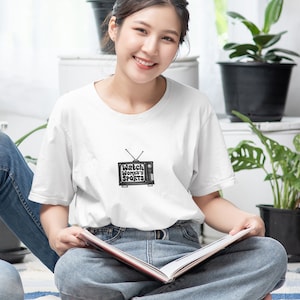 May include: A woman wearing a white t-shirt with a black and white illustration of a television with the text "Watch Women's Sports" on it. She is sitting on a striped rug and holding a book.