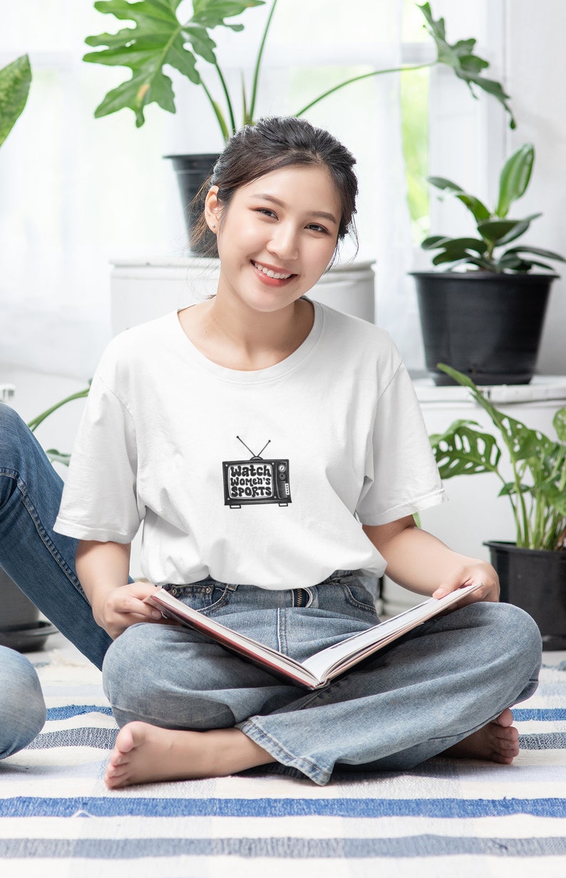 May include: A woman wearing a white t-shirt with a black and white illustration of a television with the text "Watch Women's Sports" on it. She is sitting on a striped rug and holding a book.