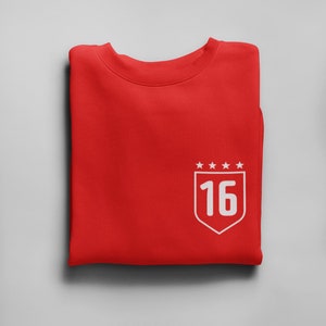 K&ouml;nnte beinhalten: Roter gefalteter Pullover mit einem wei&szlig;en gestickten Schild mit der Nummer 16 und f&uuml;nf Sternen dar&uuml;ber.