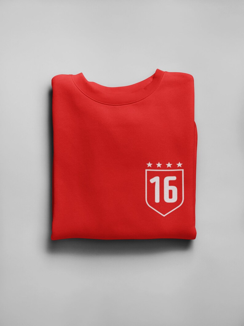 K&ouml;nnte beinhalten: Roter gefalteter Pullover mit einem wei&szlig;en gestickten Schild mit der Nummer 16 und f&uuml;nf Sternen dar&uuml;ber.