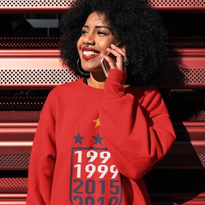 Puede incluir: Una mujer con una sudadera roja con un gr&aacute;fico azul y blanco de estrellas y los a&ntilde;os 199, 1999, 2015 y 2019. Ella sonr&iacute;e y habla por tel&eacute;fono.