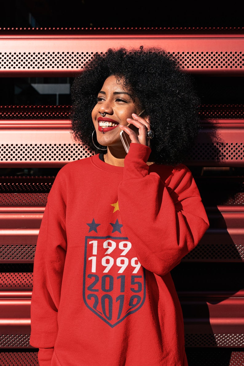 Puede incluir: Una mujer con una sudadera roja con un gr&aacute;fico azul y blanco de estrellas y los a&ntilde;os 199, 1999, 2015 y 2019. Ella sonr&iacute;e y habla por tel&eacute;fono.