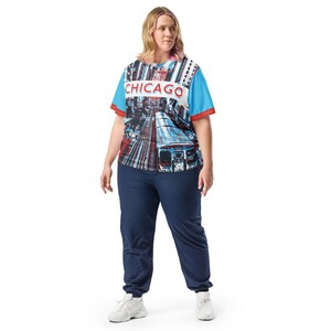 Op de afbeelding: Een vrouw draagt een blauw en rood T-shirt met een grafische print van een stadsbeeld en het woord "CHICAGO" op de voorkant. Ze draagt ook een donkerblauwe broek en witte sneakers.