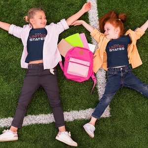 Op de afbeelding: Twee kinderen liggen op hun rug op een groen grasveld met een witte lijn die er doorheen loopt. Ze dragen blauwe shirts met de tekst "UNITED STATES" erop gedrukt. Het meisje links draagt een roze shirt over haar blauwe shirt en het meisje rechts draagt een geel shirt over haar blauwe shirt. Tussen hen in ligt een roze rugzak.