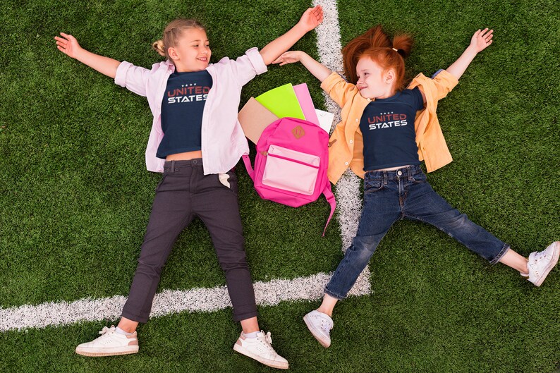 Op de afbeelding: Twee kinderen liggen op hun rug op een groen grasveld met een witte lijn die er doorheen loopt. Ze dragen blauwe shirts met de tekst "UNITED STATES" erop gedrukt. Het meisje links draagt een roze shirt over haar blauwe shirt en het meisje rechts draagt een geel shirt over haar blauwe shirt. Tussen hen in ligt een roze rugzak.