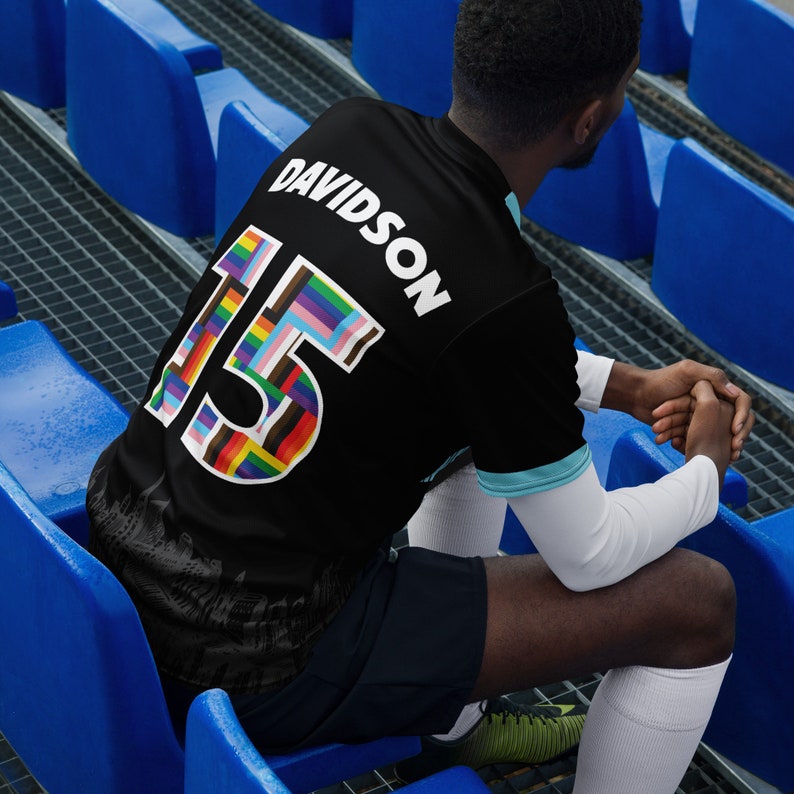K&ouml;nnte beinhalten: Ein schwarzes Fu&szlig;balltrikot mit dem Namen "DAVIDSON" und der Nummer "15" in Regenbogenfarben auf der R&uuml;ckseite. Das Trikot wird von einer Person getragen, die auf blauen Stadionb&auml;nken sitzt.
