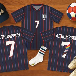 Puede incluir: Una camiseta de fútbol azul marino con rayas rojas y azules, el número 7 y el nombre A. Thompson. Una gorra de béisbol azul marino con "USA", un balón de fútbol, calcetines y tacos también están en la imagen. La camiseta tiene el logo de USA.