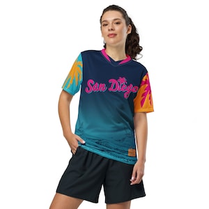 May include: A blue, teal, and orange jersey with a white v-neck collar. The jersey has a graphic print of palm trees and the text "San Diego" in pink. The jersey is being worn by a person with a ponytail.