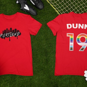 May include: Red soccer jersey with a black and white city skyline graphic on the front and the name "DUNN" and the number "19" with a rainbow outline on the back.
