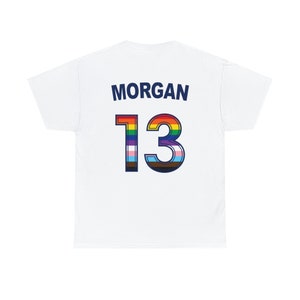 May include: White t-shirt with the name "MORGAN" and the number "13" printed on the back. The number is outlined in blue and filled with a rainbow and transgender pride flag.