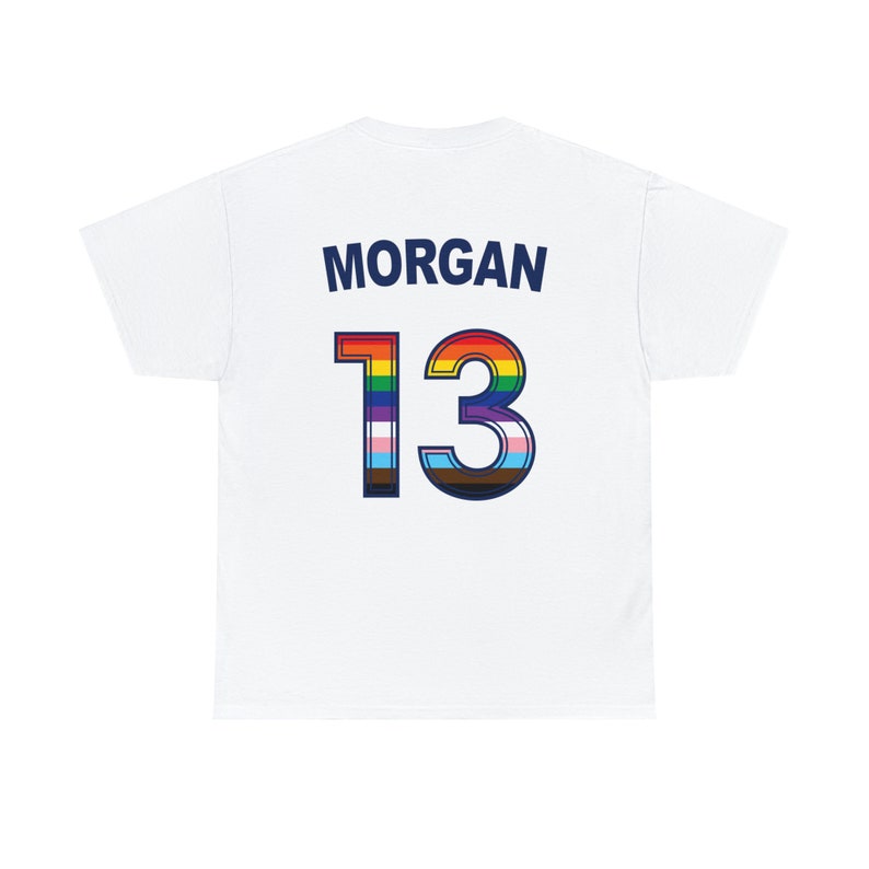 May include: White t-shirt with the name "MORGAN" and the number "13" printed on the back. The number is outlined in blue and filled with a rainbow and transgender pride flag.