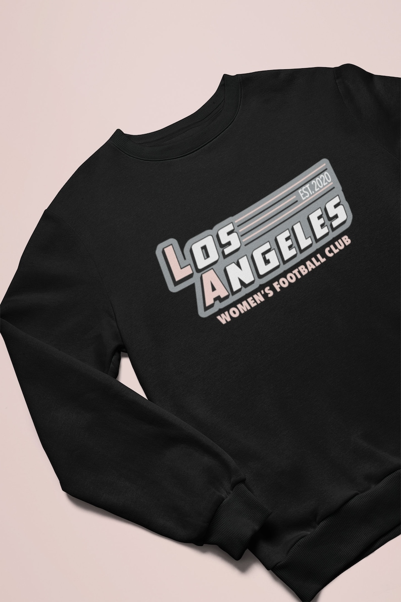 Pu&ograve; includere: Felpa nera con la scritta "LOS ANGELES WOMEN'S FOOTBALL CLUB" in bianco, grigio e rosa. Il testo "EST. 2020" &egrave; sopra la parola "LOS ANGELES". La felpa ha un girocollo e maniche lunghe.