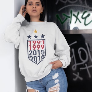 Puede incluir: Sudadera blanca con un gr&aacute;fico de escudo azul marino y rojo con los a&ntilde;os 1991, 1999, 2015 y 2019. Tres estrellas doradas est&aacute;n por encima del escudo.