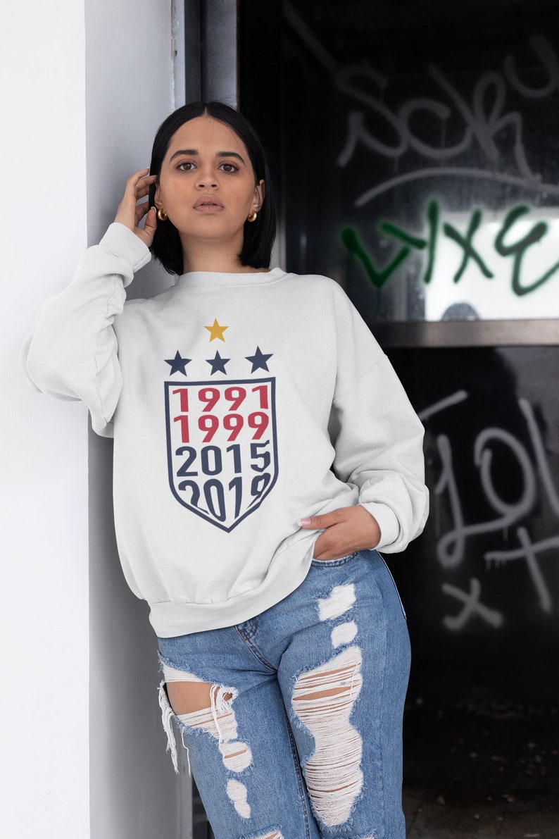 Puede incluir: Sudadera blanca con un gr&aacute;fico de escudo azul marino y rojo con los a&ntilde;os 1991, 1999, 2015 y 2019. Tres estrellas doradas est&aacute;n por encima del escudo.