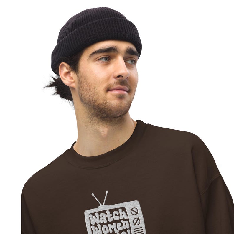 May include: A brown sweatshirt featuring a vintage television graphic with the text "Watch Women" on it. The person wearing the sweatshirt is also wearing a black beanie.