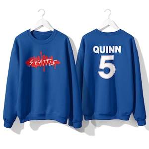 May include: Two royal blue sweatshirts. One sweatshirt has a red graphic of the Seattle skyline with the word "Seattle" written in red. The other sweatshirt has the name "Quinn" and the number "5" in white on the back.