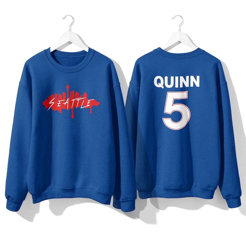 May include: Two royal blue sweatshirts. One sweatshirt has a red graphic of the Seattle skyline with the word "Seattle" written in red. The other sweatshirt has the name "Quinn" and the number "5" in white on the back.