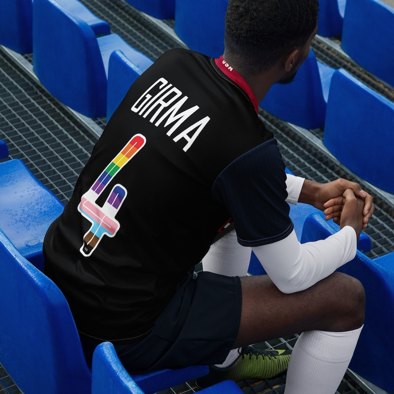 May include: A black soccer jersey with a rainbow number "4" and the name "GIRMA" on the back. The jersey is worn by a person sitting on blue stadium bleachers.