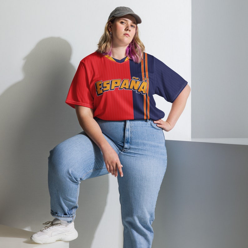 May include: A person wearing a red and blue jersey with the word "ESPA&Ntilde;A" printed on the front. The person is wearing blue jeans and a baseball cap.