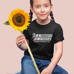 Pu&ograve; includere: Una persona indossa una maglietta nera con la scritta "LOS ANGELES WOMEN'S FOOTBALL CLUB EST. 2020" in bianco, rosa e grigio. La persona tiene in mano un grande girasole giallo e indossa jeans blu e scarpe a pois.