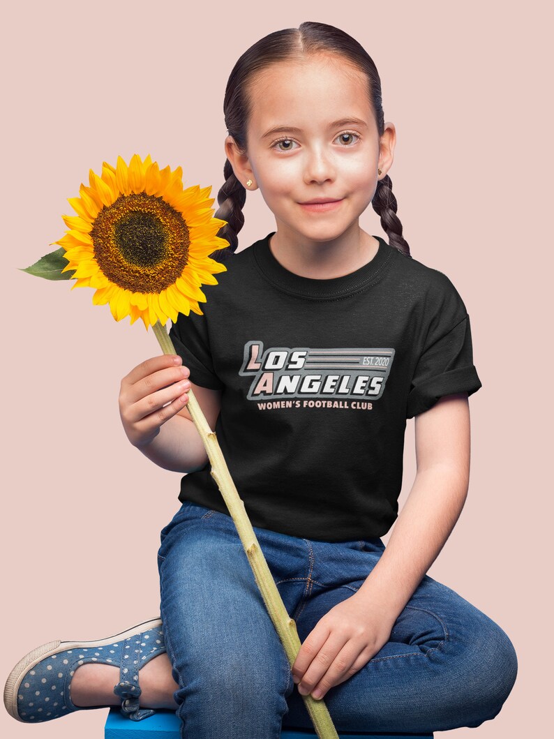 Pu&ograve; includere: Una persona indossa una maglietta nera con la scritta "LOS ANGELES WOMEN'S FOOTBALL CLUB EST. 2020" in bianco, rosa e grigio. La persona tiene in mano un grande girasole giallo e indossa jeans blu e scarpe a pois.