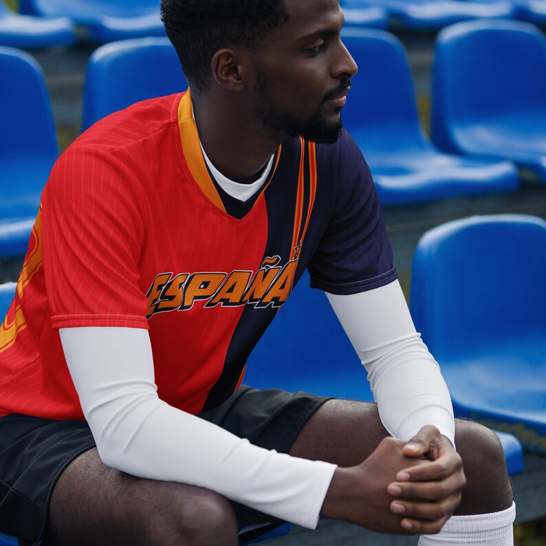 May include: A person wearing a red, black, and orange soccer jersey with the word "ESPA&Ntilde;A" printed on the front. The jersey has long white sleeves. The person is sitting on a blue bleacher.