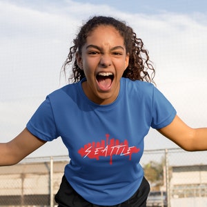 Puede incluir: Una persona con una camiseta azul con un gr&aacute;fico rojo del horizonte de Seattle. La persona est&aacute; gritando y tiene el pelo casta&ntilde;o rizado.