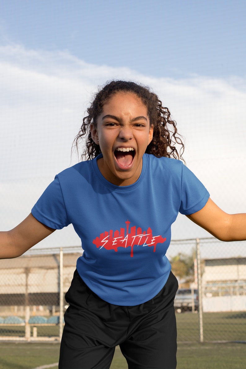 Puede incluir: Una persona con una camiseta azul con un gr&aacute;fico rojo del horizonte de Seattle. La persona est&aacute; gritando y tiene el pelo casta&ntilde;o rizado.