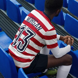 May include: A red and white striped soccer jersey with the number "13" and the name "Morgan" on the back. The jersey is worn by a person sitting on a blue bleacher.
