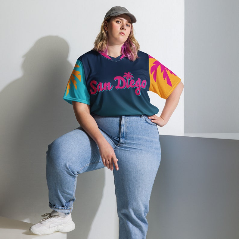 May include: A woman wearing a blue, yellow, and pink baseball jersey with the text "San Diego" in pink. She is wearing blue jeans and a grey baseball cap.