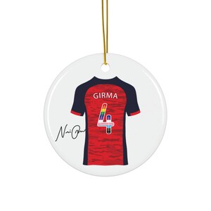 Op de afbeelding: Ronde, witte keramische kerstversiering met een voetbalshirtontwerp. Het shirt is rood met marineblauwe mouwen, de naam "GIRMA" en het nummer "4" in regenboogkleuren. Een gouden draad is bevestigd om op te hangen.