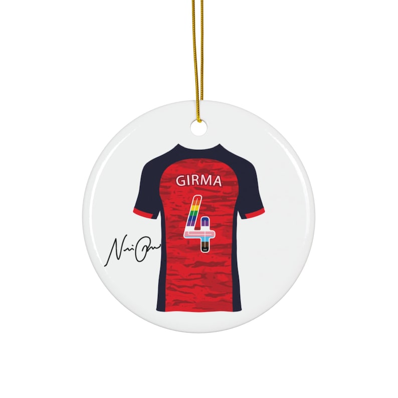 Op de afbeelding: Ronde, witte keramische kerstversiering met een voetbalshirtontwerp. Het shirt is rood met marineblauwe mouwen, de naam "GIRMA" en het nummer "4" in regenboogkleuren. Een gouden draad is bevestigd om op te hangen.