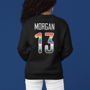 Puede incluir: Sudadera negra con el nombre "MORGAN" y el n&uacute;mero "13" en colores arco&iacute;ris en la espalda.