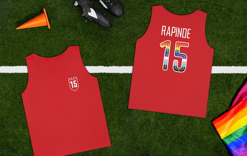 Puede incluir: Dos camisetas de tirantes rojas con el n&uacute;mero 15. Una camiseta tiene el nombre "RAPINOE" en la espalda y el n&uacute;mero 15 est&aacute; decorado con colores arco&iacute;ris. La otra camiseta tiene el n&uacute;mero 15 en un escudo blanco con estrellas.