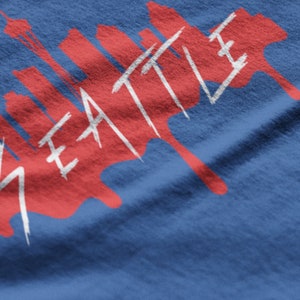 May include: A red and white graphic design on a blue t-shirt featuring the word "SEATTLE" and a skyline silhouette.