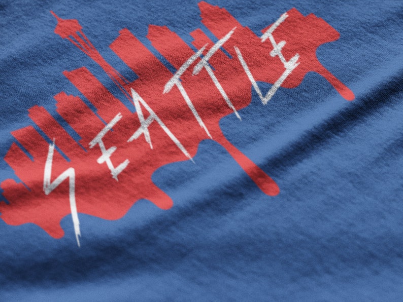 May include: A red and white graphic design on a blue t-shirt featuring the word "SEATTLE" and a skyline silhouette.