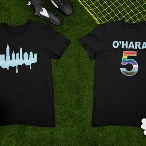 Puede incluir: Camiseta negra con un gr&aacute;fico de horizonte azul en la parte delantera y el texto "O'HARA 5" con un n&uacute;mero arco&iacute;ris en la parte posterior. La camiseta est&aacute; colocada sobre una superficie verde con un bal&oacute;n de f&uacute;tbol y un par de guantes de f&uacute;tbol.
