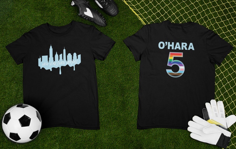 Puede incluir: Camiseta negra con un gr&aacute;fico de horizonte azul en la parte delantera y el texto "O'HARA 5" con un n&uacute;mero arco&iacute;ris en la parte posterior. La camiseta est&aacute; colocada sobre una superficie verde con un bal&oacute;n de f&uacute;tbol y un par de guantes de f&uacute;tbol.