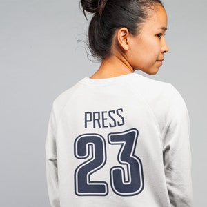 K&ouml;nnte beinhalten: Ein wei&szlig;er Pullover mit einer dunkelblauen Nummer 23 und dem Wort "PRESS" auf der R&uuml;ckseite.