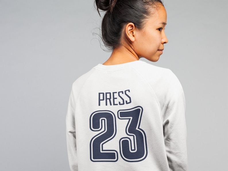 K&ouml;nnte beinhalten: Ein wei&szlig;er Pullover mit einer dunkelblauen Nummer 23 und dem Wort "PRESS" auf der R&uuml;ckseite.