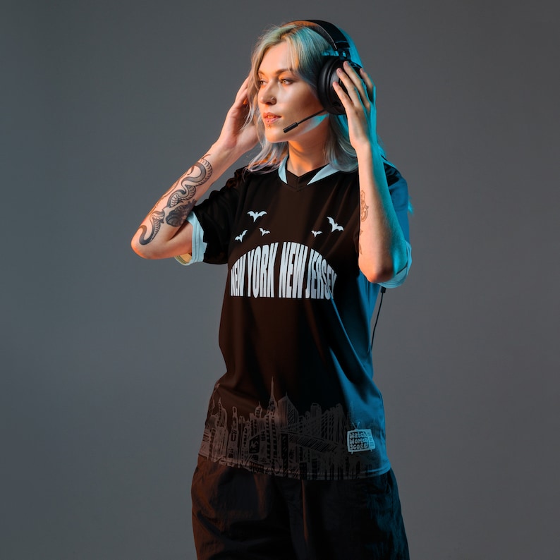 K&ouml;nnte beinhalten: Eine Frau tr&auml;gt ein schwarzes T-Shirt mit einem wei&szlig;en Aufdruck, der "New York New Jersey" und ein Stadtbild zeigt. Sie tr&auml;gt eine schwarze Hose und hat ein Tattoo auf ihrem Arm. Sie tr&auml;gt Kopfh&ouml;rer mit einem Mikrofon.