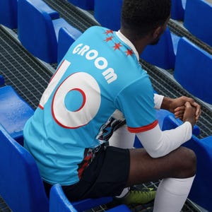 Op de afbeelding: Een blauw shirt met het woord "groom" en het nummer "10" op de rug. Het shirt heeft een wit en rood sterrendesign op de voorkant.
