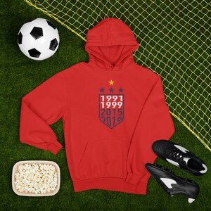 May include: Red hooded sweatshirt with a navy blue shield design featuring the years 1991, 1999, 2015, and 2019.