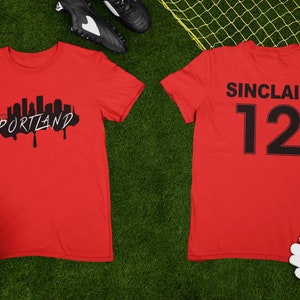 May include: Two red t-shirts on green grass. One shirt features a black Portland skyline graphic with the city name in white. The other shirt displays "SINCLAIR" and the number "12" in black. A soccer ball, cleats, and gloves are also visible.