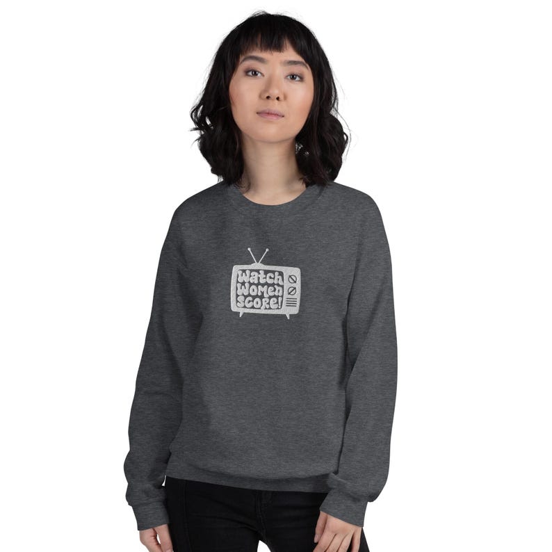 May include: A dark grey sweatshirt featuring a white graphic of a vintage television screen with the text "Watch Women Score!" inside.